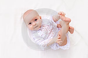 A small baby girl 6 months old is lying on a light bed in a beautiful white bodysuit and smiling at the camera, the baby is lying