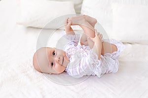 A small baby girl 6 months old is lying on a light bed in a beautiful white bodysuit and smiling at the camera, the baby is lying