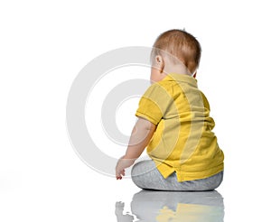 Small baby in a yellow polo sits next to us on the floor, turning his head, looking at a white background.