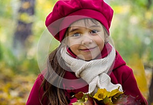 A small beautiful girl with big eyes smiling in warm autumn, wearing a pink beret and a coat with dry leaves in hands.