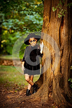 a small, beautiful girl in a black dress and hat, a face in Halloween makeup, stands near a tree