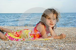 A small, beautiful girl with curly hair lies on the beach on the sand. Close-up and with place for text.