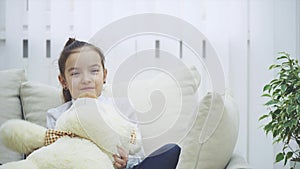 Small beautiful kid sits hugging big teddy-bear, talking to the camera.