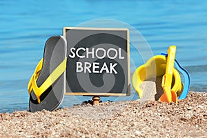 Small black chalkboard with text School Break, beach toys and flip flops on sand near sea. Seasonal holidays