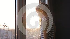 Small Blond Baby Kid Stands At Window Looks At The Street Against Sunset