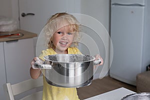 Small blond curly girl in the kitchen holding big metal pot with daugh. Girl is happily smiling, flour is on her hands