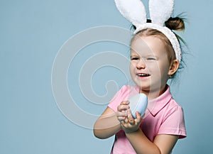 Small blond girl in pink cosy home clothing and white decorative fur ears standing, holding blue egg in hands and smiling
