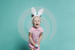 Small blond girl in pink cosy home dress and white decorative fur ears standing, holding blue egg in hands and feeling cheerful