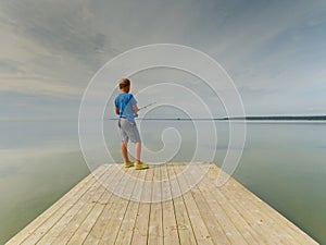 Small blond hair boy is fishing at the end of wooden mole. Smooth water level in bay
