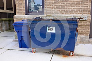 Small blue dented dumpster on wheels on a sidewalk next to a blue trash can in front of a brown brick wall