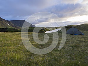 Small blue tent standing alone on green grass on mossed creek banks in Hornstrandir Iceland, snow patched hills and cliffs,