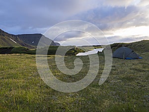 Small blue tent standing alone on green grass on mossed creek banks in Hornstrandir Iceland, snow patched hills and cliffs, photo