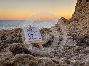 Small board with "Katie, will you marry me?" written on it on the rocky shore during sunset photo