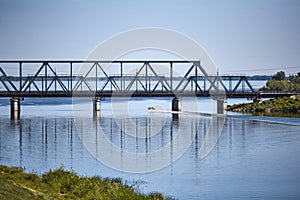 A small boat sails along the river in a summer sunny day under a railway bridge