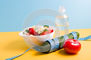 small bottle of water, salad in a white bowl, blue measuring tape around a cucumber,