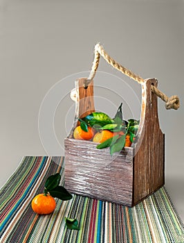 A small box with handles filled with tangerines. On tangerines leaves. A box is standing on a colorful napkin