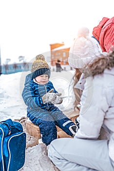A small boy of 4-6 years old, sitting on a bench, in the winter in the city on the rink. Waiting for Mom to put on