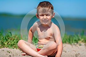 A small boy with a bare torso in shorts sits on the sand on the lake at noon