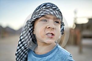 Small boy in checkered keffiyeh grimacing outdoors