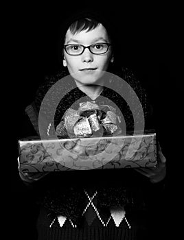 Small boy or cute nerd kid in glasses, hat and fashionable knitted scarf on black background holds red present box with