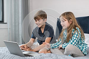 Small Boy And Girl Use Laptop Computer Sitting On Bed In Bedroom, Brother And Sister Surfing Internet