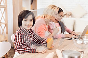 Small boy is holding glass of orange juice while sitting with his grandmother and sister in kitchen.