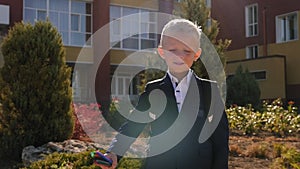 A small boy in a jacket with a backpack holds markers near the school building.