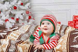 Newborn baby girl dressed in gnome costume lying on white fur carpet among christmas decorations.