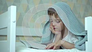 A small boy reads a book lying under a blanket