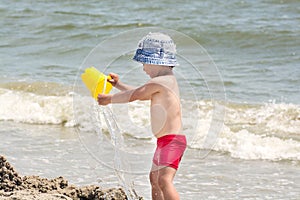 A small boy on the seashore pours water from a bucket against the background of the sea and waves,