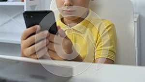 Small boy sitting in front of laptop, watching video on smartphone, addiction