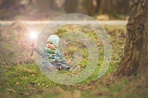 Small boy is sitting on grass and handling magic wand in spring park near big tree