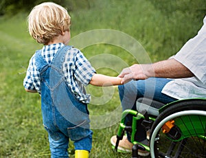 Small boy with unrecognizable senior grandfather in wheelchair on a walk on meadow in nature.