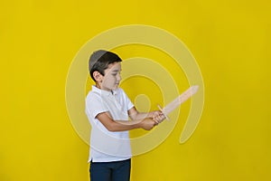 A small boy in a white shirt stands on a yellow background.In the Hands of a child a toy wooden sword
