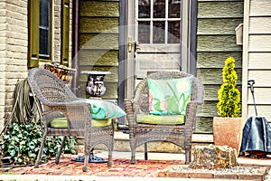 Small brick patio on brick and rustic wood siding house with two wicker chairs with bright pillows and plants and water hose hangi