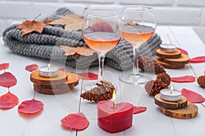 Small burning candles, two glasses with rose wine, cones, dry red leaves, a gray scarf knitted on a white wooden table.