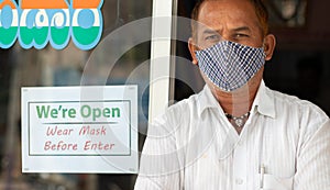 Small business owner in medical mask standing in front of door with we are open wear mask notice board - concept of support local