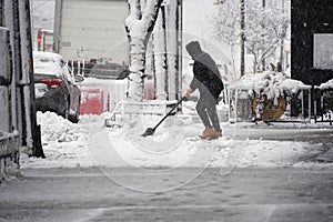 Small business owner outside store shoveling ice and slush on downtown city street with falling snow