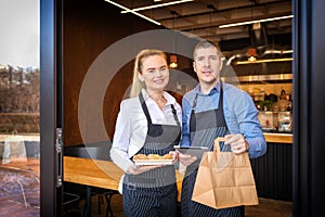 Small business owners couple standing in doorway of trendy restaurant delivering takeaway orders and attending customers