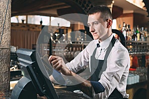 Small business, people and service concept - happy man or waiter in apron at counter with cashbox working at bar or