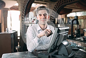 Small business, people and service concept - happy man or waiter in apron at counter with cashbox working at bar or