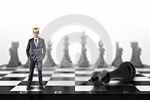 A small businessman with a golden crown on his head stands on a chessboard near a fallen black king.