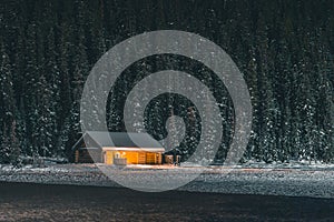 Small cabin on the banks of frozen Lake Louise during late winter night with snow capped trees around and in the background