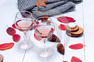 Small candles, two glasses with rose wine, cones, dry red leaves, gray scarf knitted on a white wooden table.