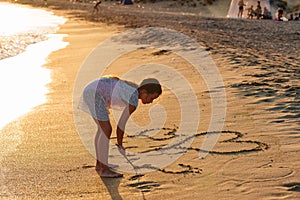 Small Caucasian girl drawing figures of hearts with a stick on sandy beach at summer sunset