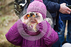 Small caucasian girl wearing a purple hat and purple coat looking through binoculars in a forest