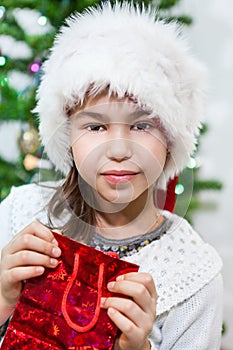 Small Caucasian girl in white fur Santa hat holding red gift bag in hands