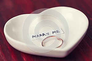 Small ceramic saucer in the shape of a heart with a ring and a note that says `Marry me`