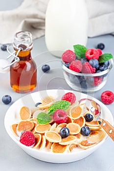 Small cereal pancakes with maple syrup, raspberry, blueberry and milk, in a white bowl, vertical