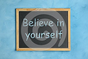 Small chalkboard with motivational quote Believe in yourself on light blue background, top view